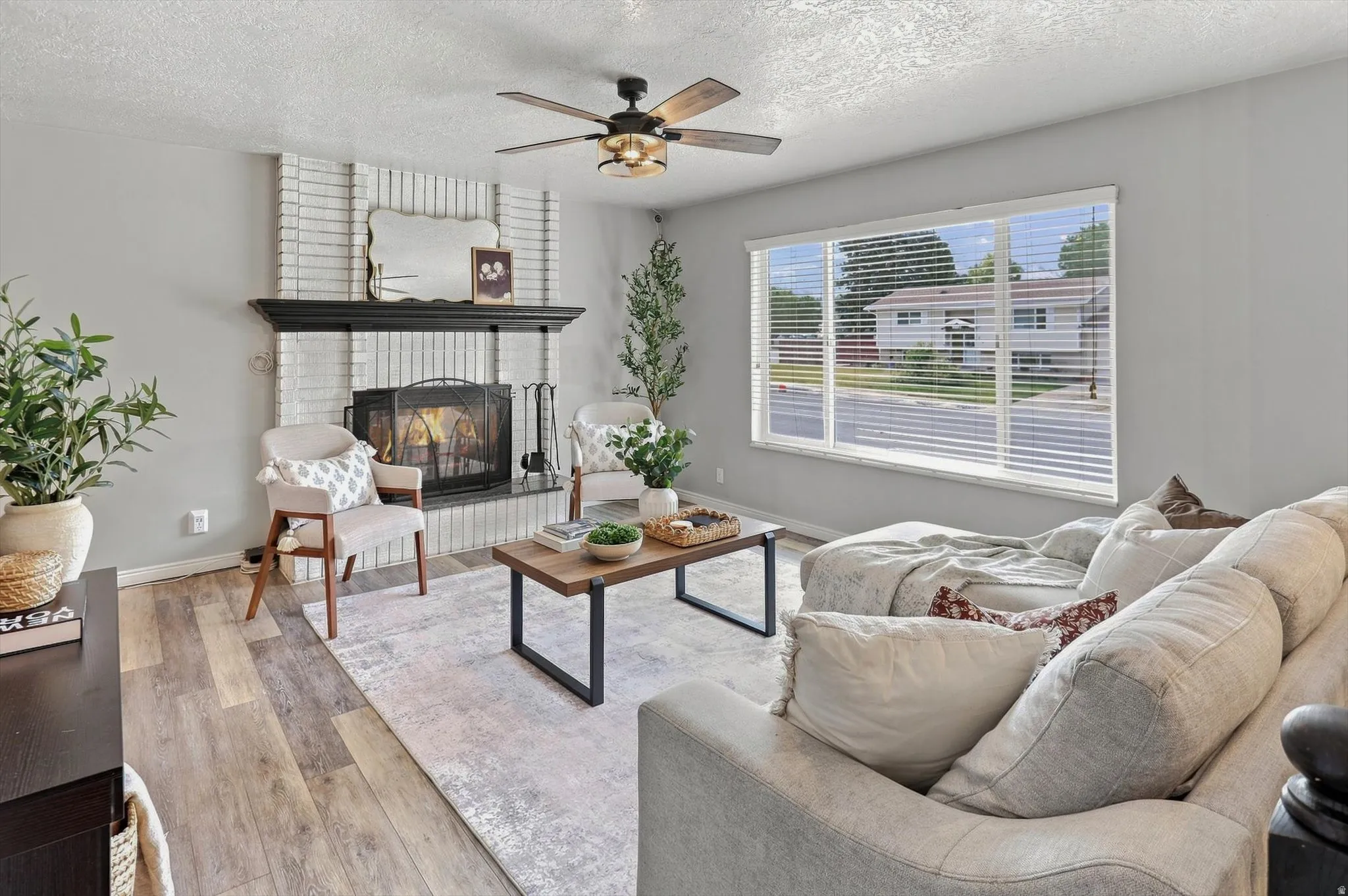 Living area featuring a white brick fireplace with a black mantel, wood-finish flooring, and a large picture window with blinds