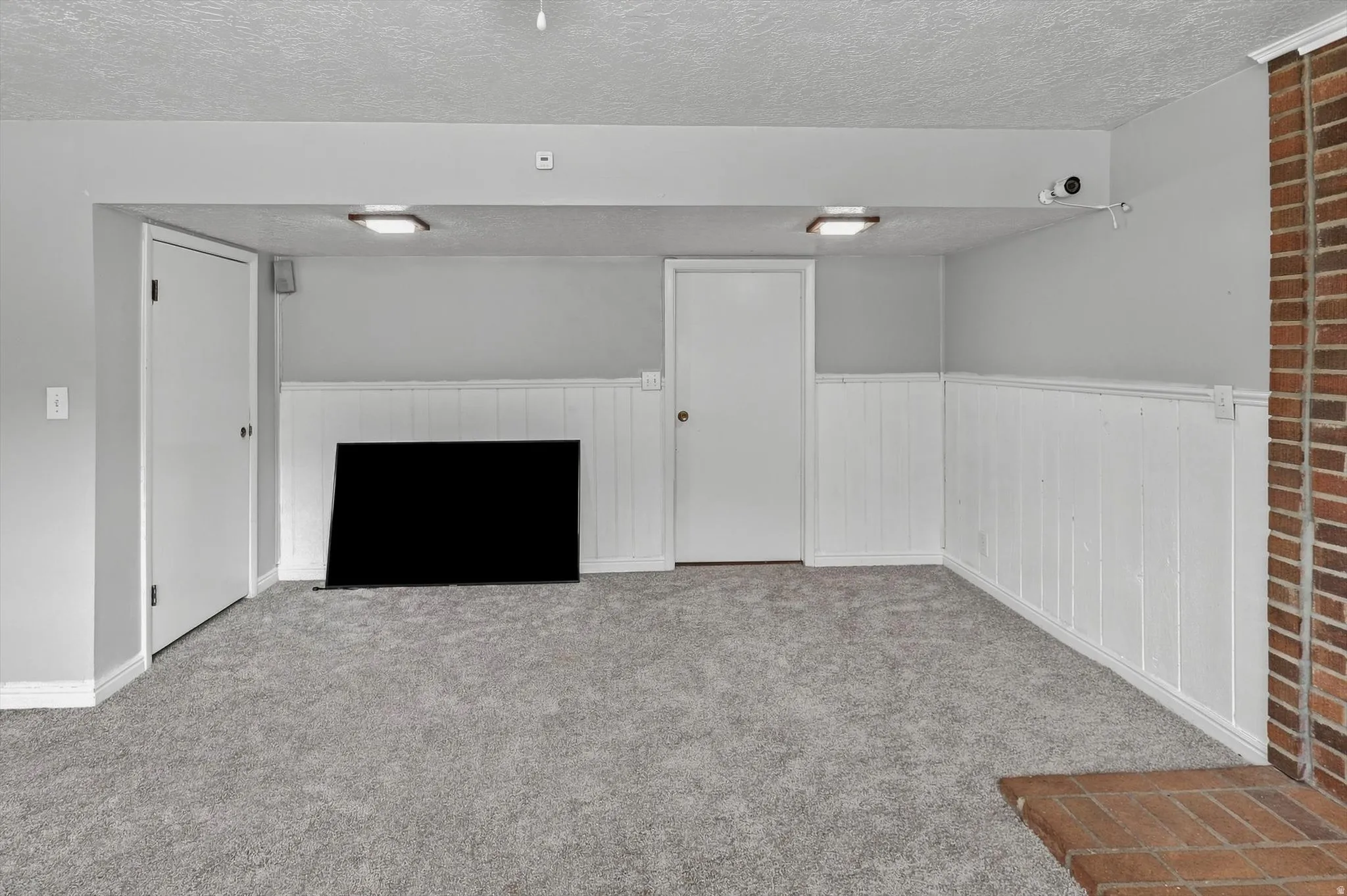 Spacious room featuring light gray carpeting, white wainscoting, and a brick fireplace surround with a hearth