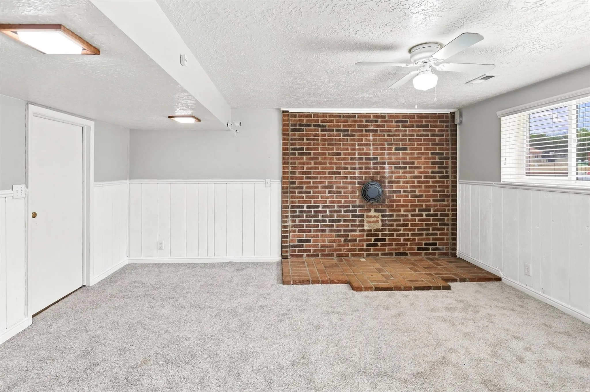 Interior space featuring a brick accent wall with a hearth, gray carpeting, white wainscoting, and a ceiling fan