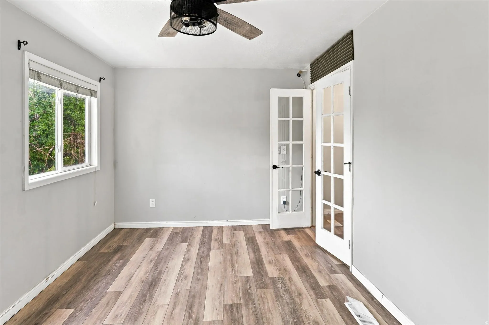 Room featuring light gray painted walls, wood-finish flooring, a window with a roller shade, and white French doors