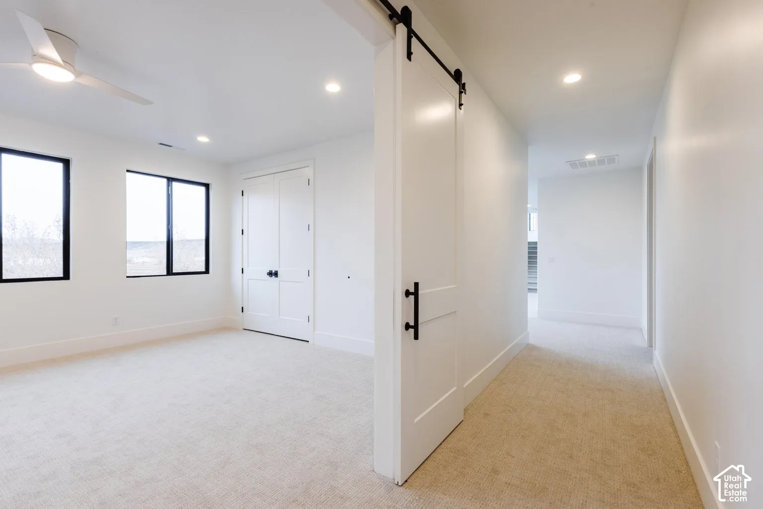 Bright room with light-toned carpeting, featuring two large windows with black frames, a double-door closet with dark hardware, and a ceiling fan with integrated lighting