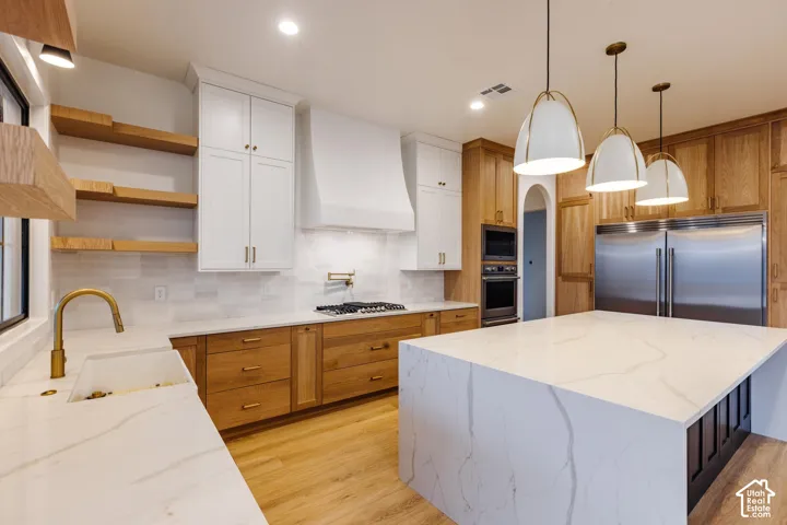 Gourmet kitchen featuring a large central island with a waterfall countertop, white upper cabinetry, natural wood-finish lower cabinetry, and integrated stainless steel appliances