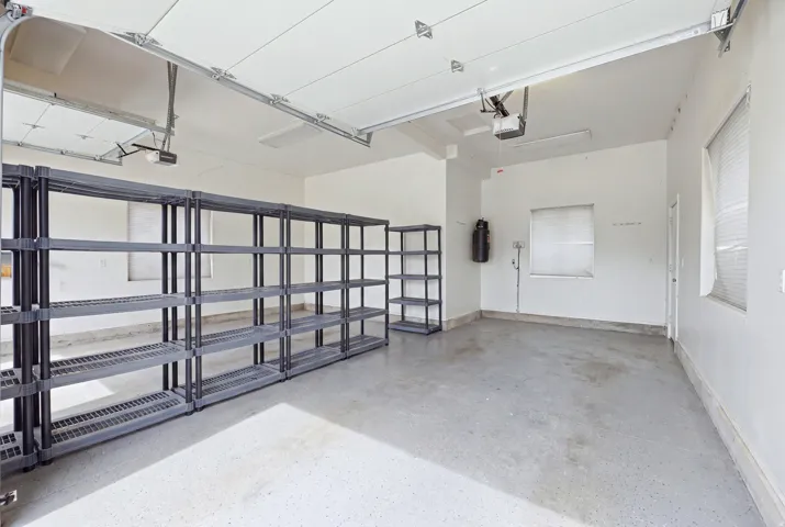 Spacious three car garage featuring ample shelving, two automatic door openers, a concrete floor, and multiple windows with horizontal blinds