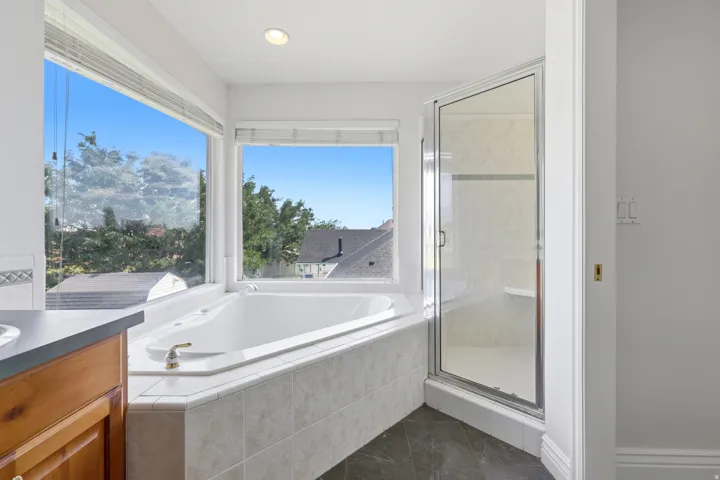 Soaking tub with tiled surround, large windows, glass-enclosed shower, wood vanity with integrated sink, and dark floor tiling