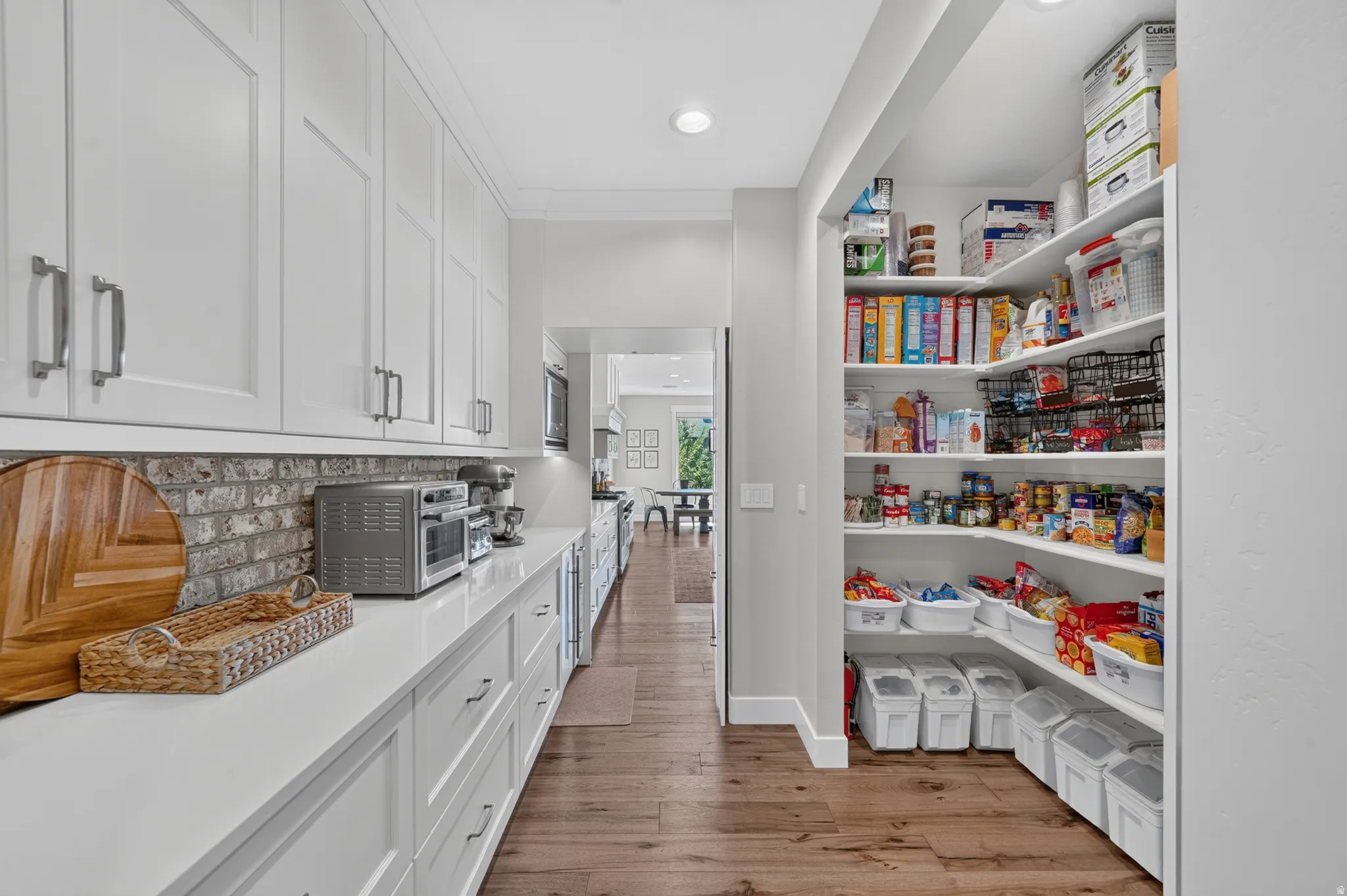 Galley-style kitchen with extensive white cabinetry, light-toned countertops, exposed brick-finish backsplash, and wood-finish flooring