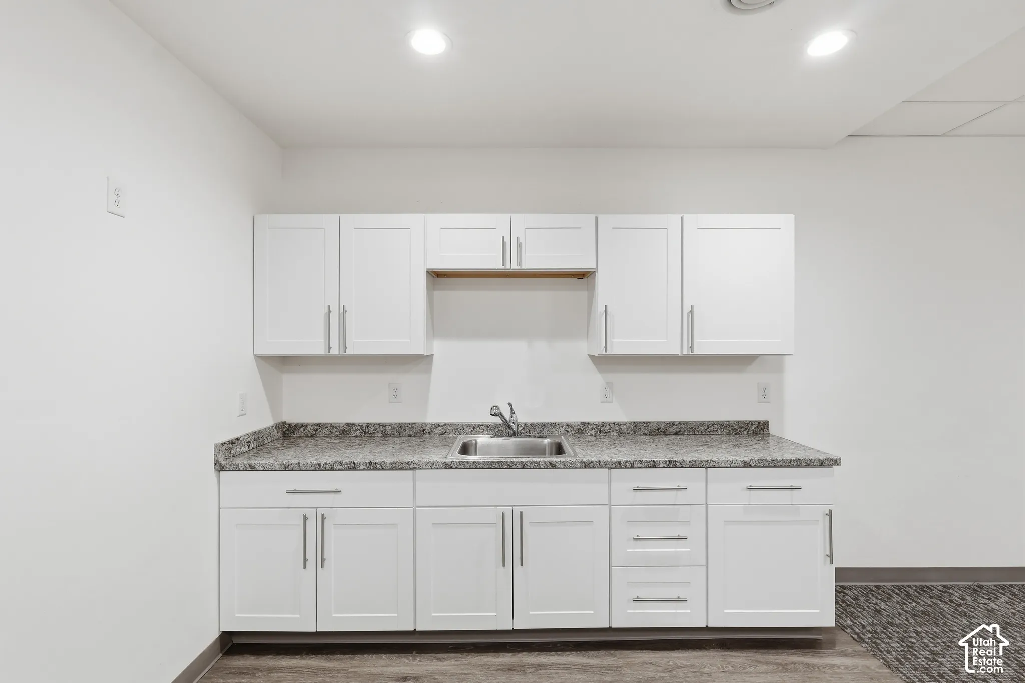 White shaker-style cabinetry with brushed nickel hardware, gray speckled countertop, stainless steel sink, and wood-finish flooring