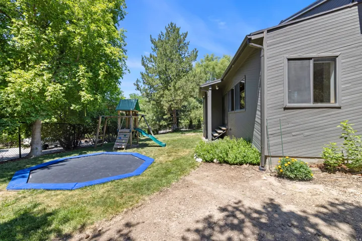 Expansive backyard featuring a manicured lawn, established trees, a wooden play structure with a slide, and with a in-ground trampoline