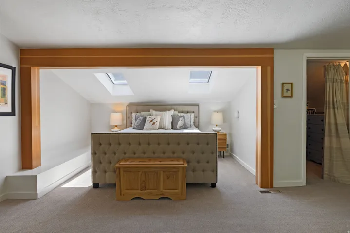 Primary bedroom featuring a tufted upholstered bed frame, two skylights, light wood architectural beams, and light-colored carpeting