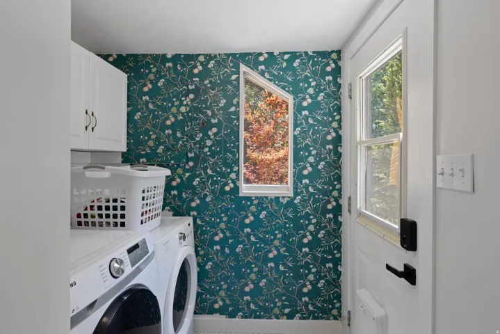 Utility room featuring a patterned accent wall, white cabinetry with dark hardware, a window offering natural light, and a white door with integrated window panels