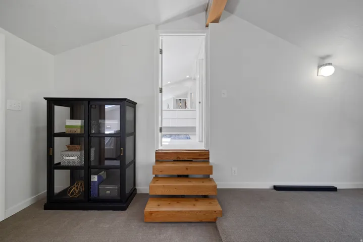 Carpeted space with light gray walls, an exposed wood beam, and a wall-mounted light fixture leading to the primary bathroom