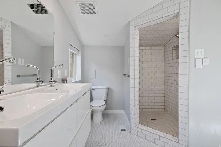 Primary modern bathroom featuring a dual-sink vanity with chrome fixtures, a tiled walk-in shower with subway tile surround, white hex tile flooring, and a window for natural light