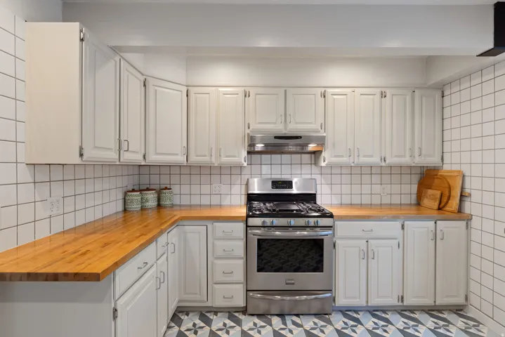 Kitchen featuring white cabinetry, butcher block countertops, white square tile backsplash, and stainless steel gas range with coordinating vent hood