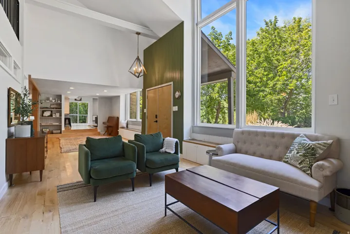 Vaulted ceilings with exposed beams, expansive floor-to-ceiling windows, light wood-finish flooring, and a contemporary pendant light fixture
