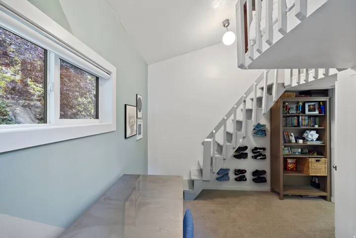 White baluster staircase with a built-in bookshelf below, light blue wall accent, and a window with a roller shade. Bed loft overhead.