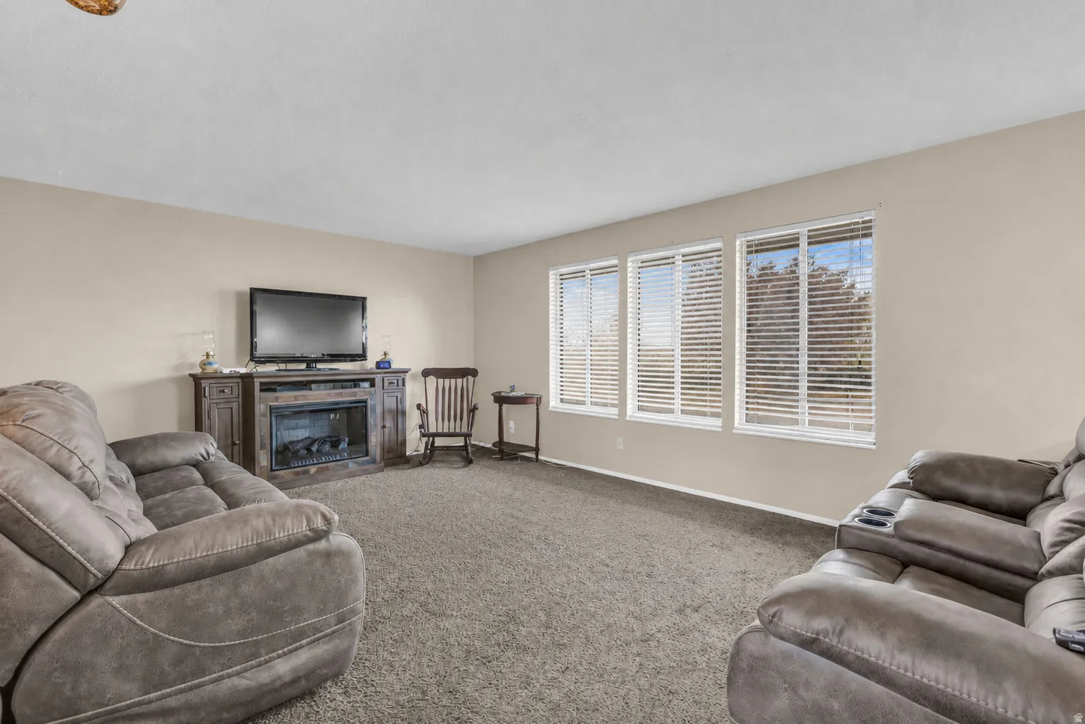 Neutral wall paint, carpet flooring, a multi-pane window with horizontal blinds, and a media console with an integrated electric fireplace