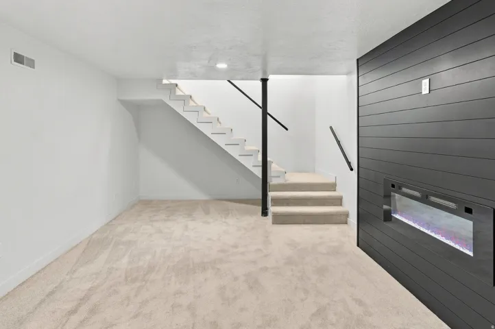 Lower level space featuring an integrated electric fireplace within a dark shiplap wall, neutral carpet flooring, and a staircase with white risers, carpeted treads, and black metal handrails