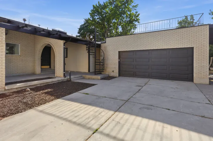 Cream brick exterior featuring an arched entryway, a dark panel garage door, a concrete driveway, a spiral staircase, and an upper-level railing