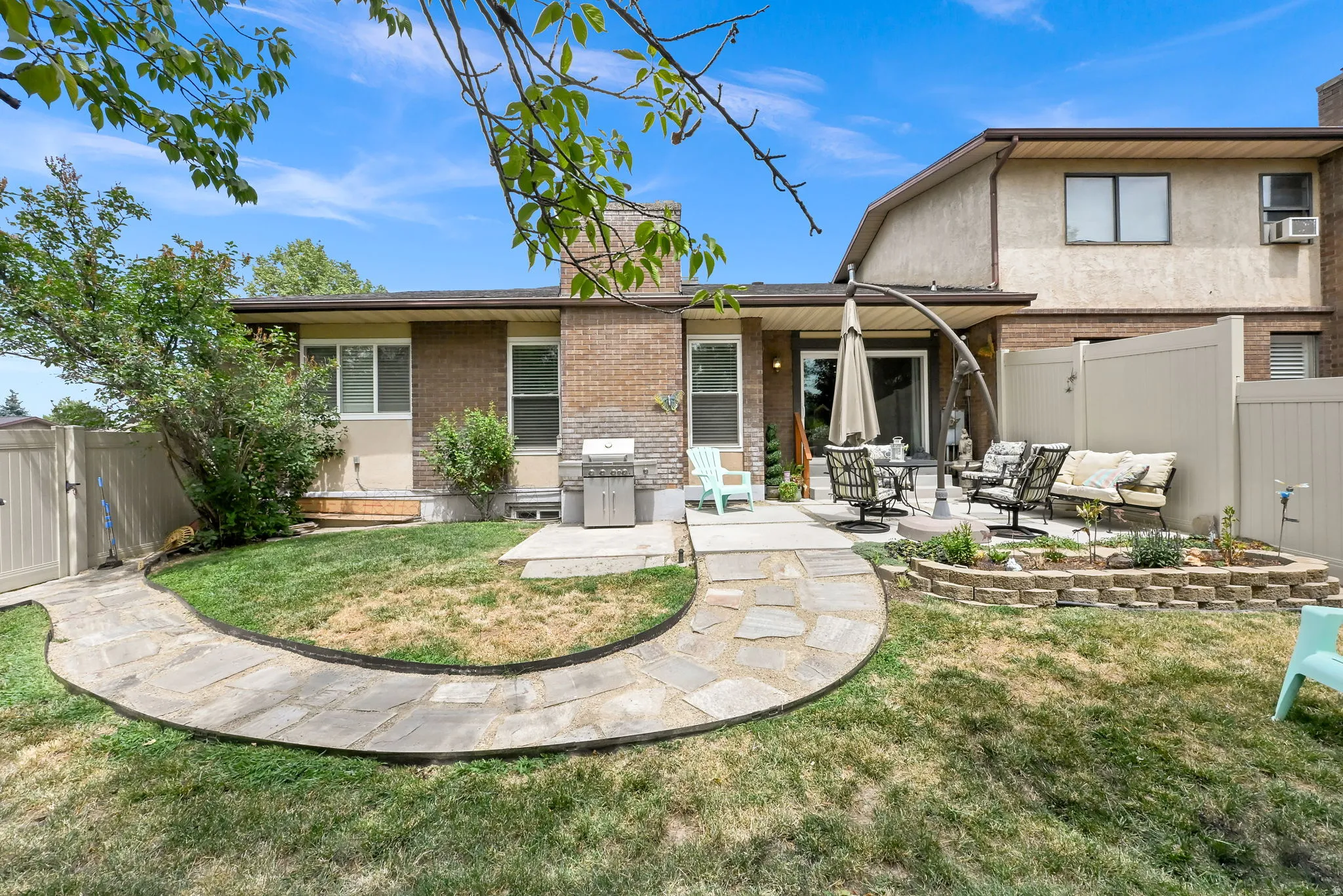 Expansive backyard featuring a stone pathway leading to a concrete patio, a raised garden bed with stone block retaining walls, a privacy fence, and mature landscaping
