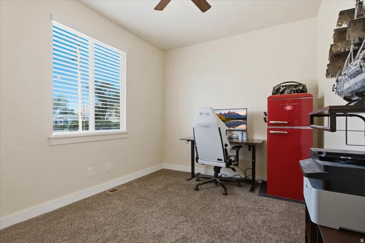 Carpeted room featuring a large window with horizontal blinds, light beige wall paint, white baseboards, and a ceiling fan with wood-finish blades
