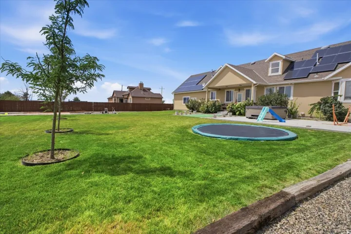 Expansive green lawn with young trees, a brown privacy fence, a paved patio, and solar panels installed on the roof of the home