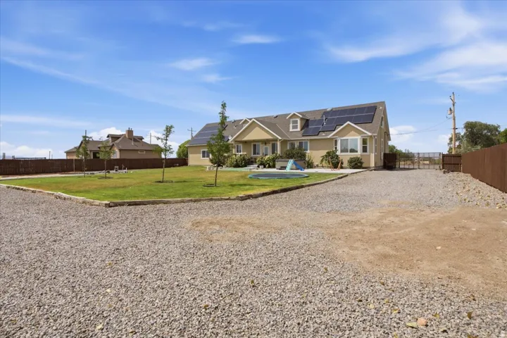 Single-story residence featuring a gravel driveway, an expansive lawn area, mature fencing, roof-mounted solar panels, and a dormer window