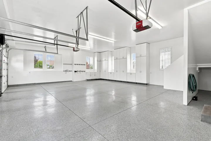 Spacious garage featuring a finished epoxy floor, extensive built-in cabinetry, multiple windows with plantation shutters, overhead fluorescent lighting, and two garage door openers