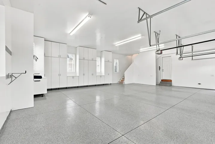 Spacious garage featuring a speckled epoxy floor, extensive built-in white cabinetry, multiple windows with white louvered shutters, and overhead fluorescent lighting