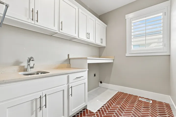 White shaker cabinetry with brushed nickel hardware, stone-finish countertops, stainless steel sink, a built-in workstation, and brick-patterned flooring