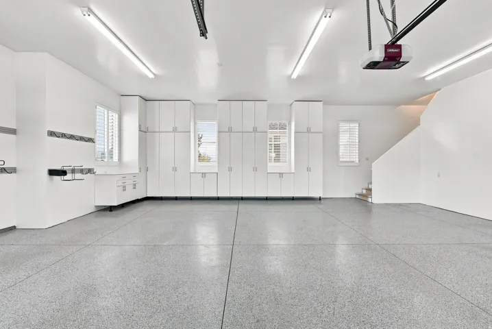 Spacious garage featuring a light gray speckled epoxy floor finish, extensive white built-in cabinetry, and multiple windows with white louvered blinds
