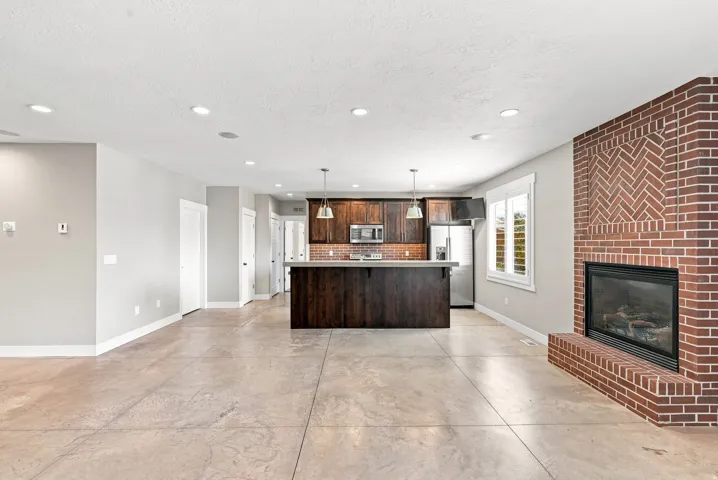 Spacious open-concept living area featuring a prominent brick fireplace with a herringbone accent, a kitchen island with pendant lighting, dark wood cabinetry, and stainless steel appliances
