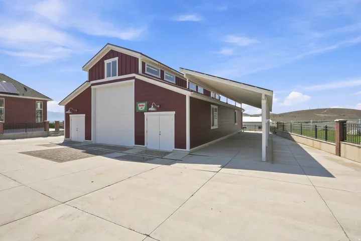 Red-sided barn-style structure featuring a large main garage door, two pedestrian doors with exterior lighting, and an extended carport
