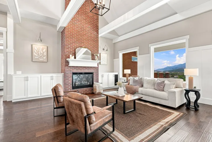 Spacious living area featuring a floor-to-ceiling brick fireplace, vaulted ceilings with exposed beams, built-in white cabinetry, dark wood-finish flooring, and large windows offering exterior mountain scenery