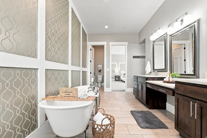 Freestanding soaking tub with chrome fixtures, dual vanities featuring dark wood cabinetry, light-toned countertops, and framed mirrors with overhead lighting