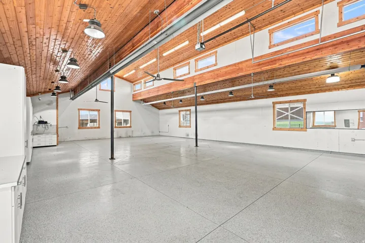 Spacious open area featuring an epoxy-coated concrete floor, exposed wood ceiling with industrial lighting, and multiple windows providing natural light