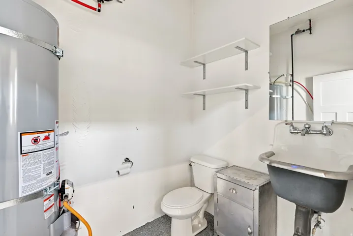 Utility area featuring a water heater, toilet, pedestal sink with wall-mounted faucet, and two wall-mounted shelves