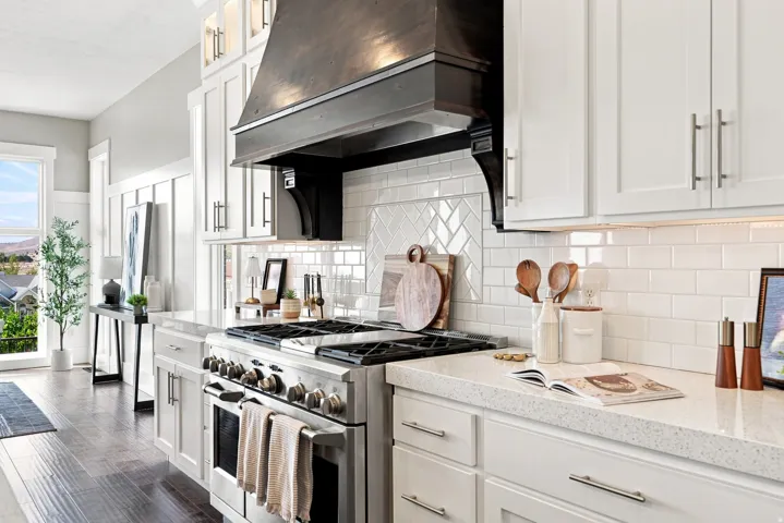 Gourmet kitchen featuring a stainless steel range with a dark wood-finish hood, white shaker cabinetry, light-toned countertops, and a subway tile backsplash with a decorative herringbone pattern