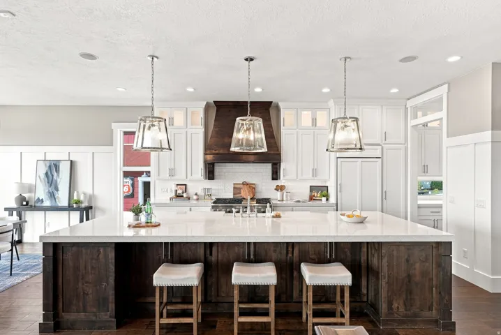 Spacious kitchen featuring a large central island with a light-toned countertop, white cabinetry with upper glass-front accents, dark wood-finish range hood, and recessed lighting
