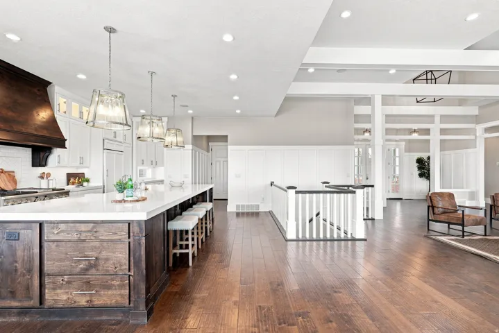 Open-concept kitchen and living area featuring dark wood-finish flooring, a kitchen island with white countertop, white cabinetry with a wood range hood, and a staircase with white balusters