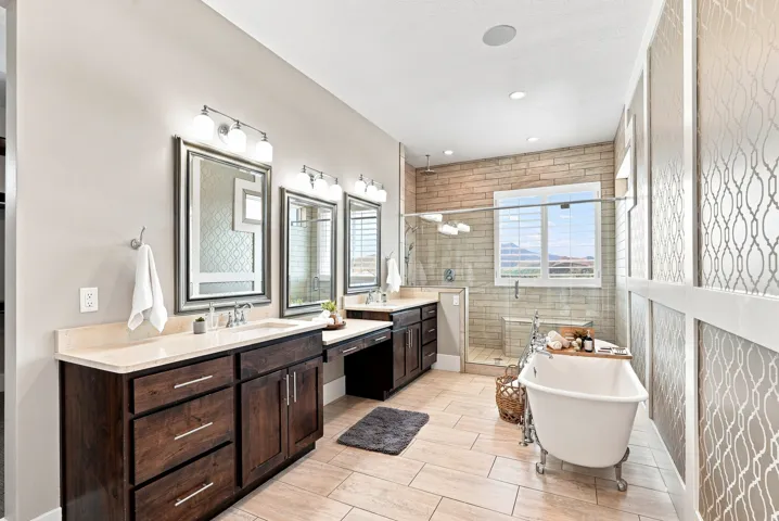 Primary bathroom featuring a freestanding clawfoot tub, dual dark wood-finish vanities with light countertops, a glass-enclosed shower with subway tile, and light-toned tile flooring