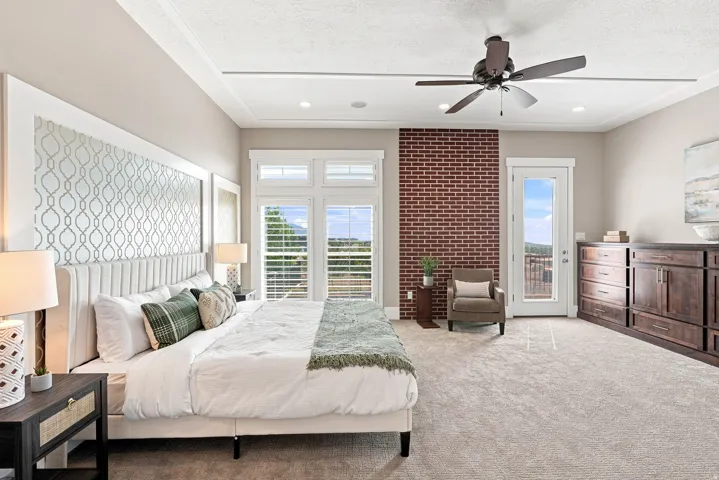 Spacious bedroom featuring light-colored carpeting, a tray ceiling with a dark-bladed ceiling fan, and a brick accent wall