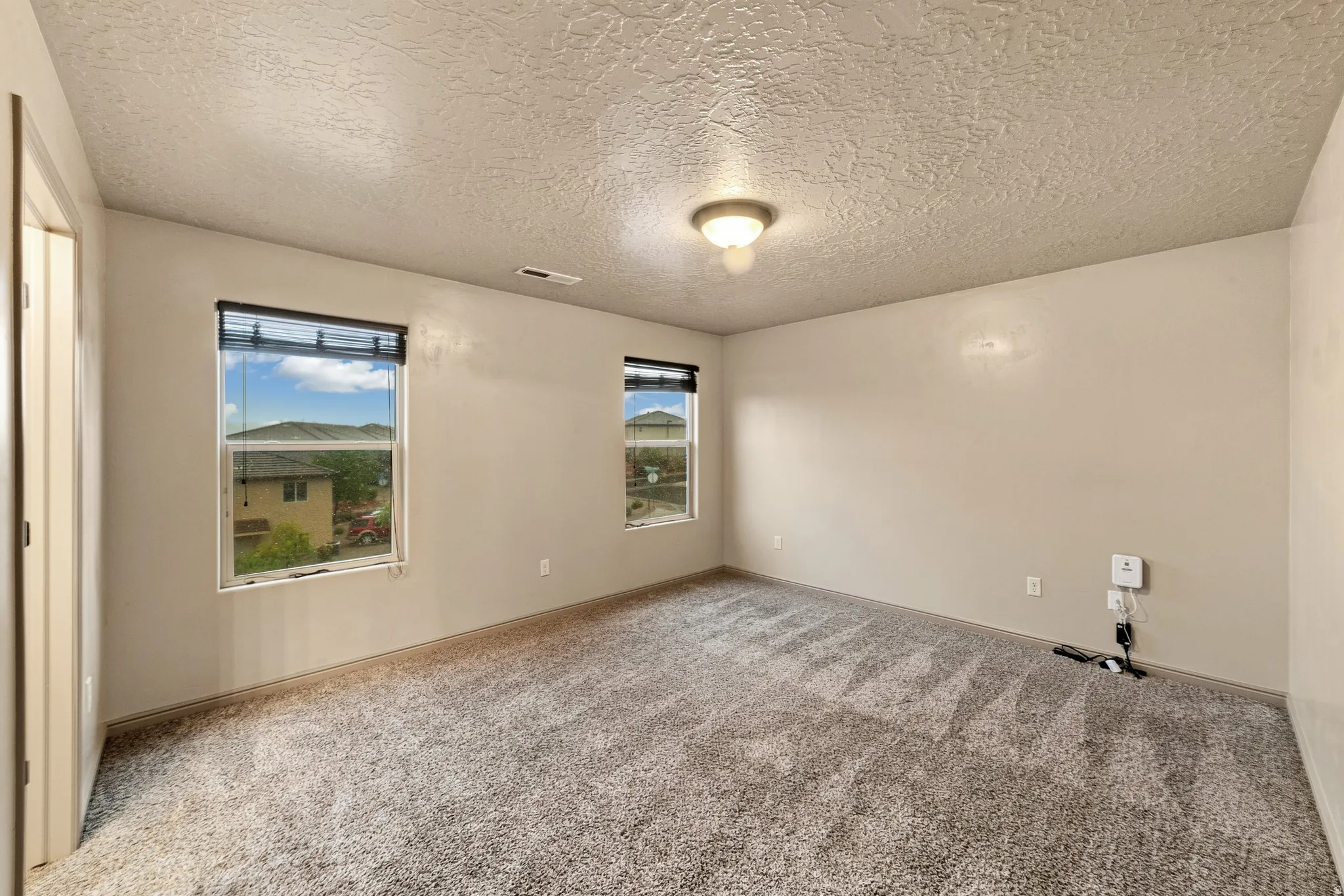 Spacious room featuring neutral carpeting, two windows with blinds, a ceiling-mounted light fixture, and textured ceilings