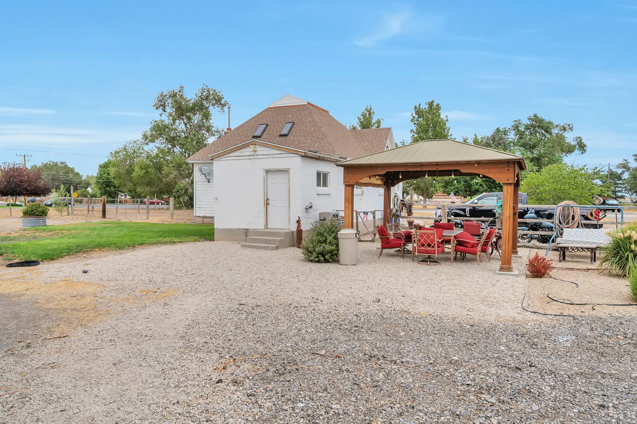 Gravel outdoor area featuring a wooden gazebo with a metal roof, an adjacent white structure with a brown shingle roof, and visible skylights