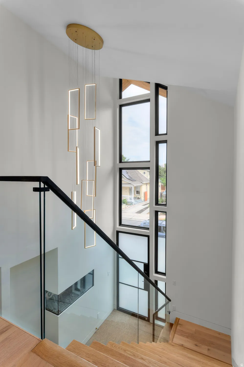 Contemporary multi-level entryway featuring a prominent linear chandelier, a multi-pane window system with black frames, hardwood stairs, and a glass-paneled railing with black metal accents