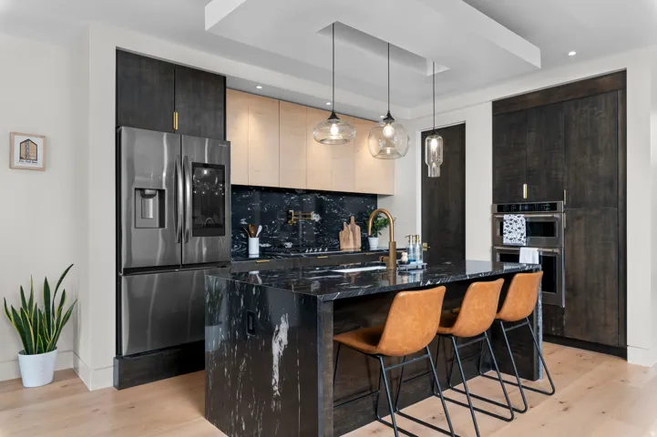 Gourmet kitchen featuring a large waterfall island with seating, stainless steel appliances, dark stone countertops and backsplash, and light wood-finish flooring