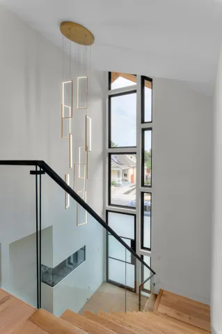 Contemporary multi-level entryway featuring a prominent linear chandelier, a multi-pane window system with black frames, hardwood stairs, and a glass-paneled railing with black metal accents