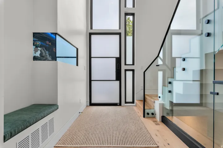 Modern entryway featuring a multi-paneled frosted glass door and surrounding windows, light wood-finish flooring, and a contemporary staircase with glass railing