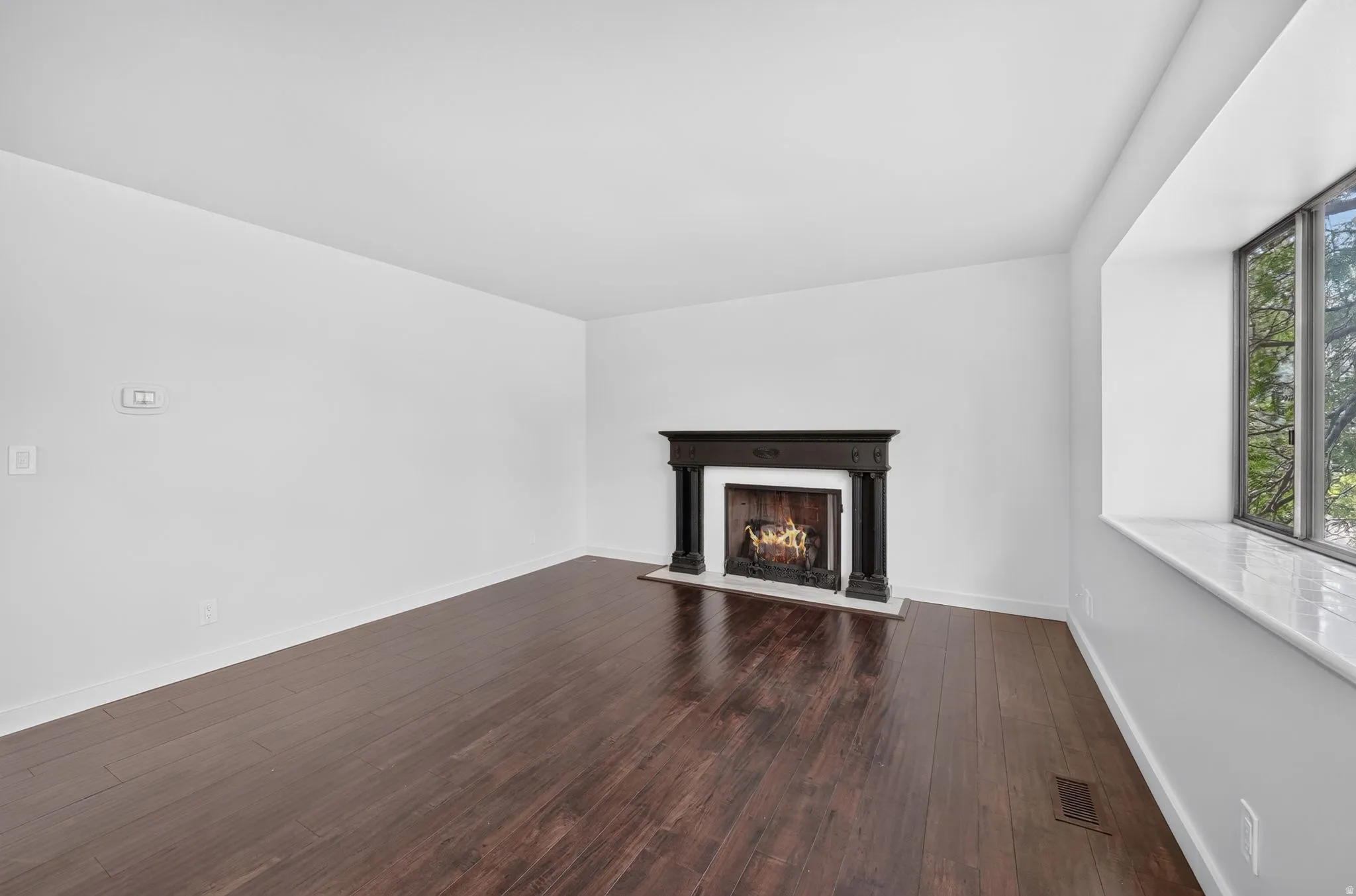Bright living space featuring dark wood-finish flooring, a decorative fireplace with a dark wood mantel, and a large bay window with a white marble sill