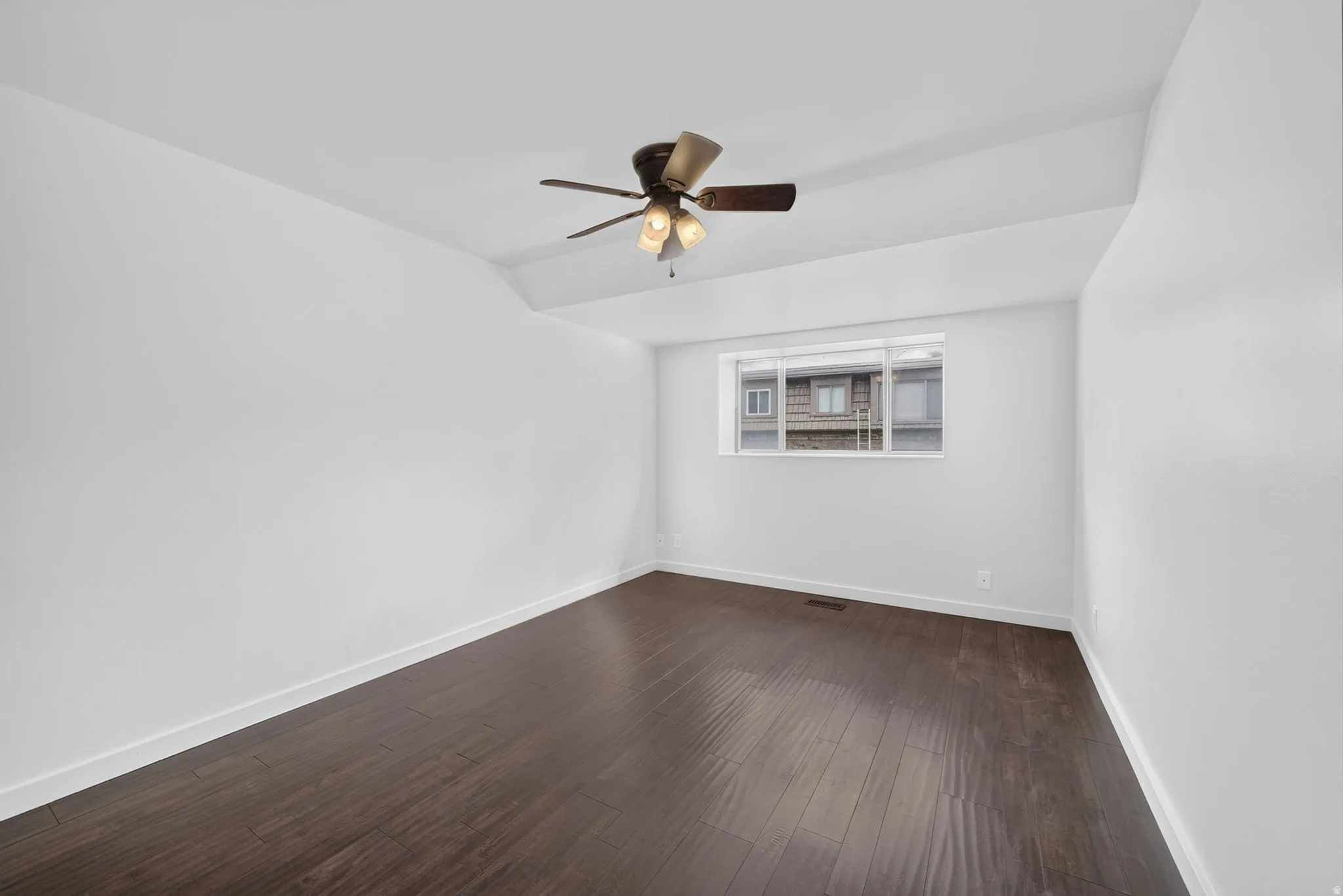 Spacious room featuring rich wood-finish flooring, a contemporary ceiling fan, white baseboards, and a large window providing ambient light