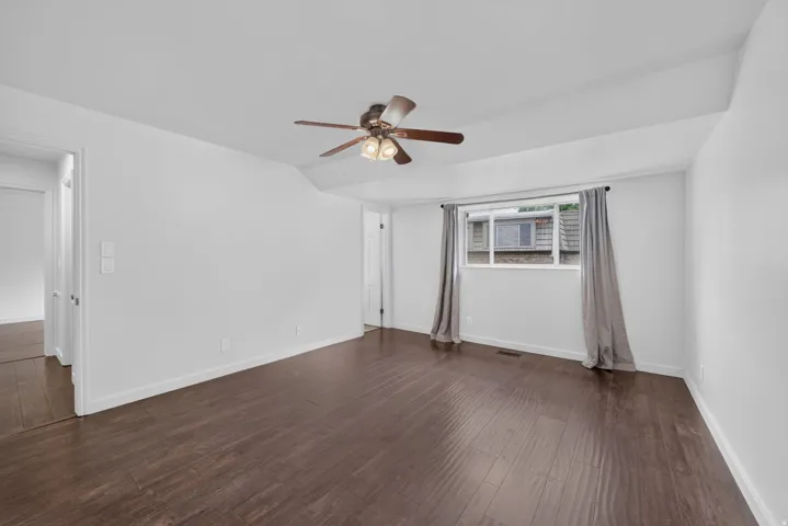 Spacious room featuring dark wood-finish flooring, white walls, and a ceiling fan with integrated lighting