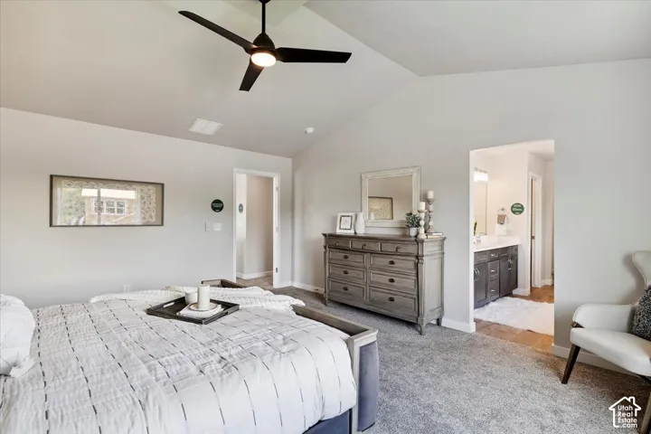Primary bedroom featuring a vaulted ceiling with a black ceiling fan, light gray carpeting, and a visible doorway to an adjoining bathroom featuring a vanity with dark wood-finish cabinetry and quartz countertops